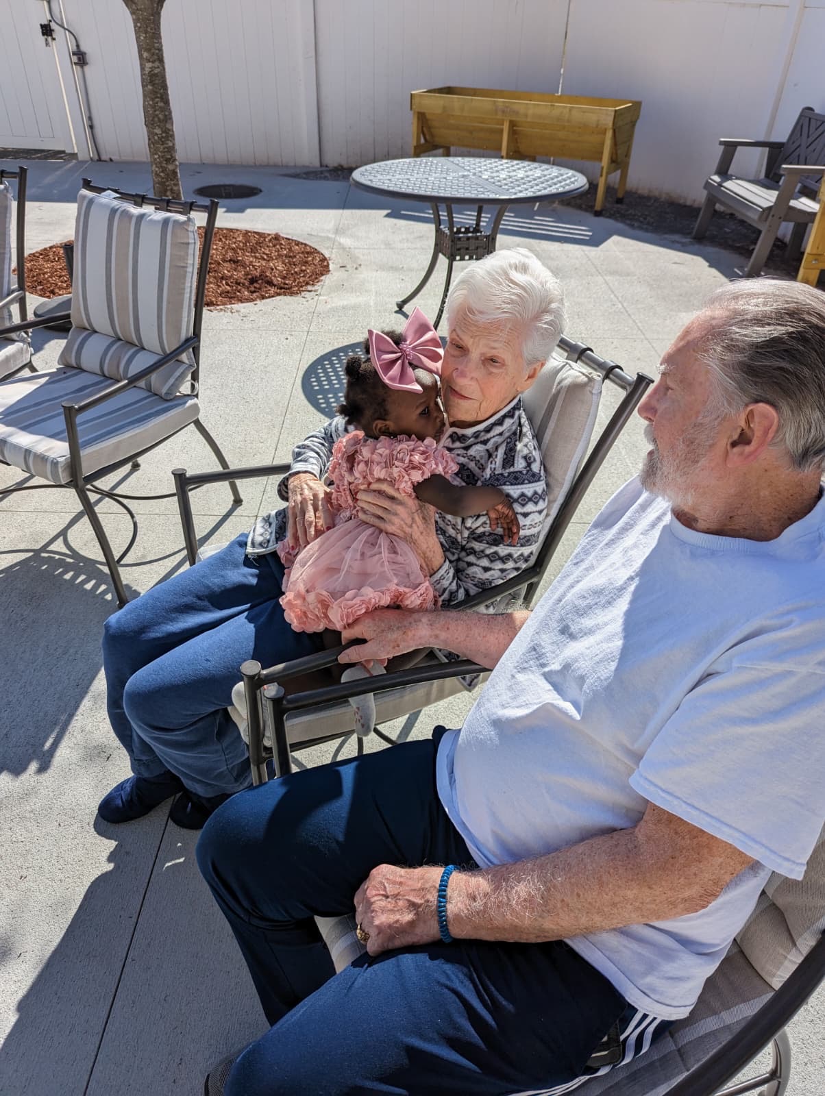An older couple sitting outside on a patio with a small child in a pink dress on the woman's lap.