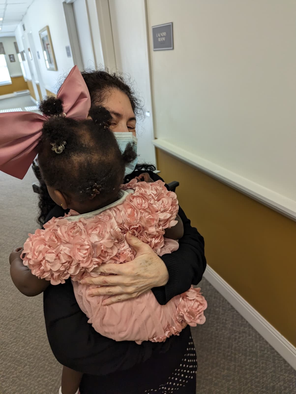 A caregiver holding and hugging a small child in a pink rose dress in a hallway.