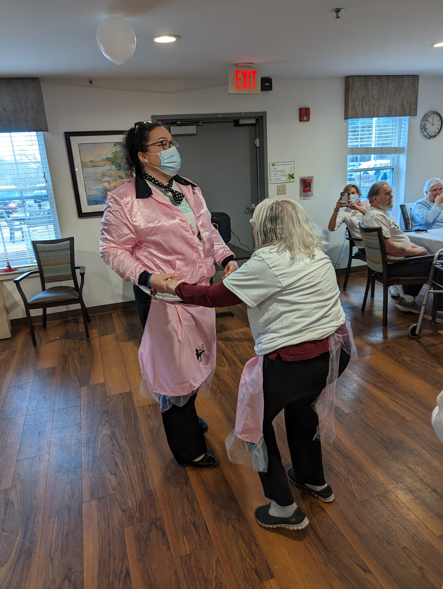A caregiver in a pink 50s-style jacket dancing with a resident at a community event.