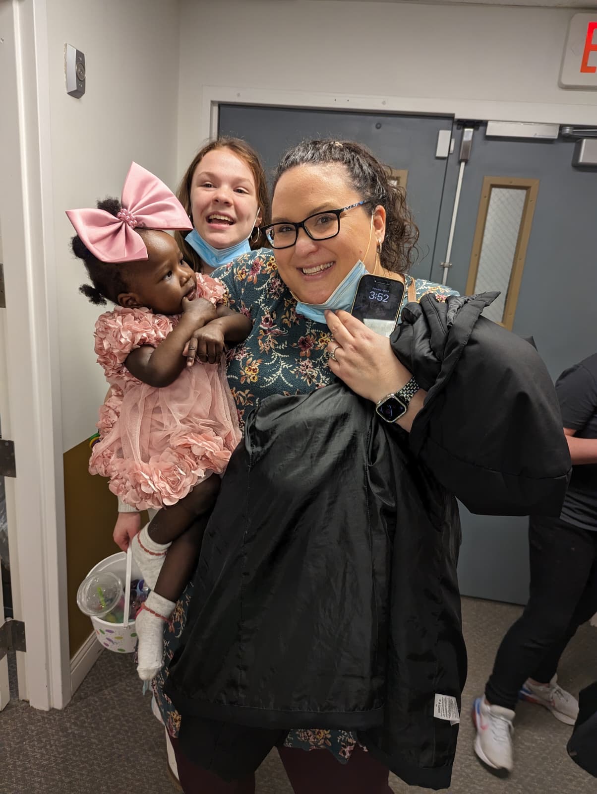Two Selena M Barach caregivers smiling with a small child in a pink dress at a community event.
